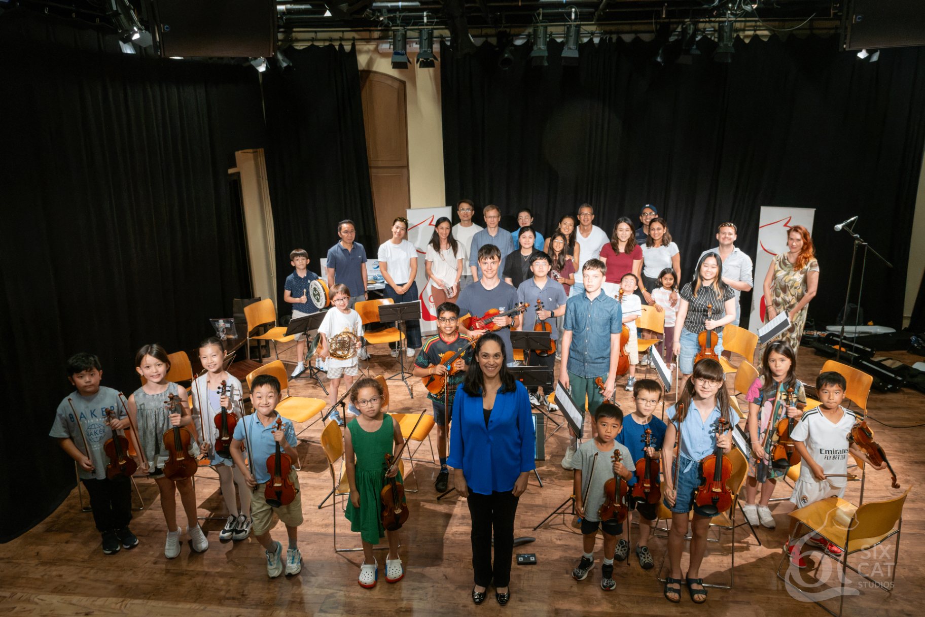 Aniela Chaudhuri stands at the front of a stage group of young violinists holding violins, with family and teachers behind them